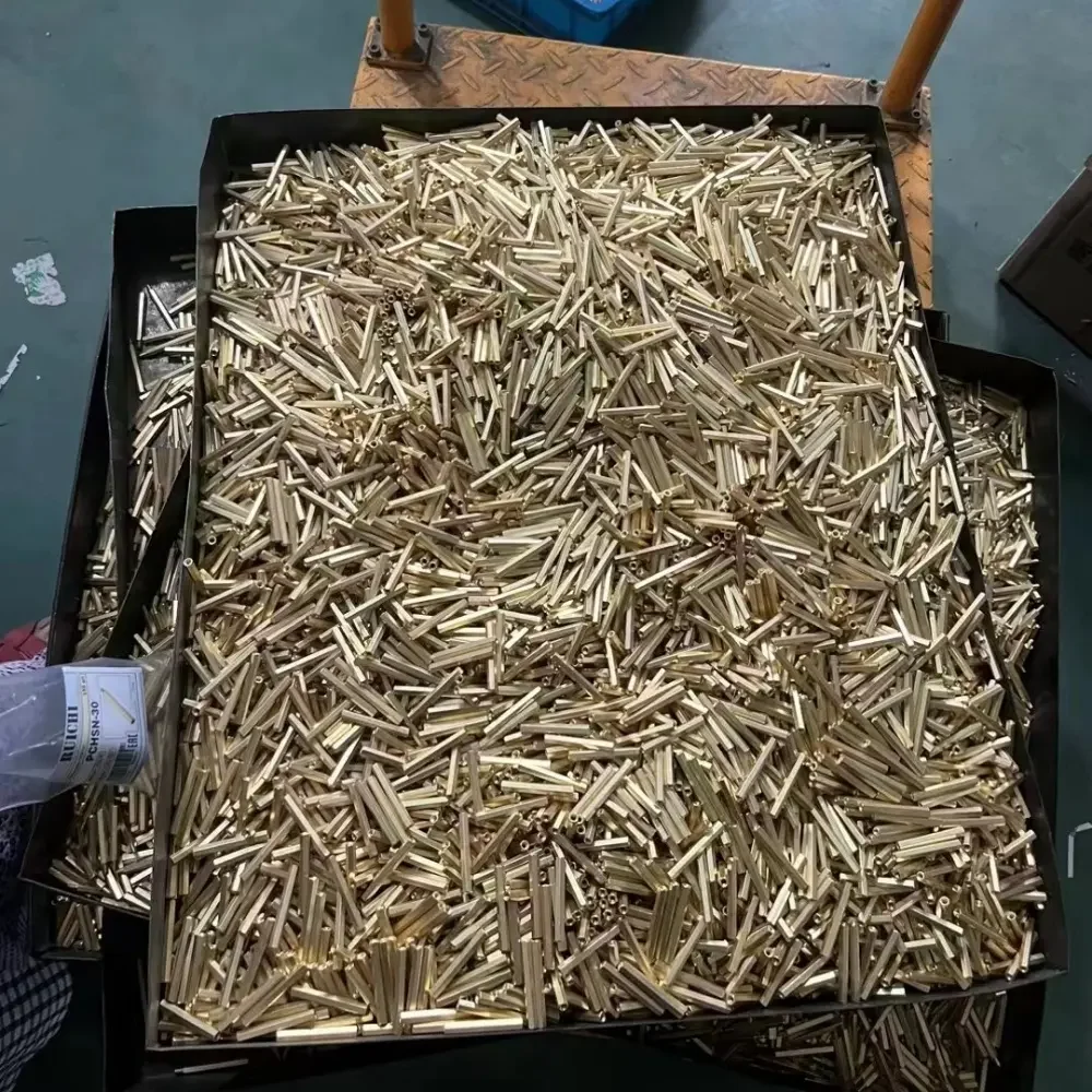 The image shows a large number of raw brass components, likely terminals for DC shunts, loosely gathered in a container. This represents an initial stage in the manufacturing process where the raw materials are prepared for further machining and assembly.